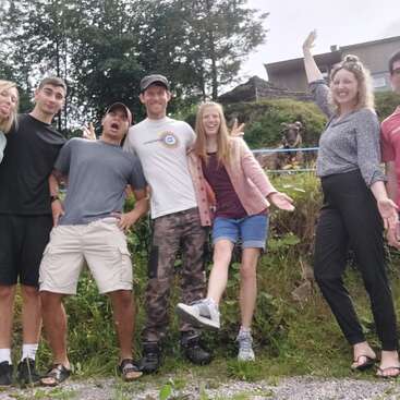 The image depicts a group of seven individuals posing for a photo, with a cow standing behind them, set against a backdrop of trees and buildings.