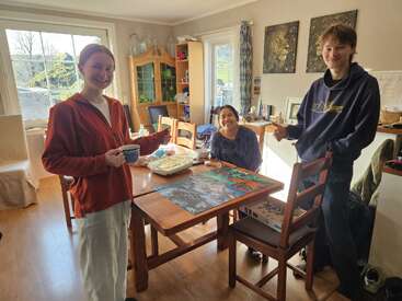 Trois personnes souriantes sont réunies autour d'une table en bois sur laquelle se trouve un puzzle coloré. La lumière du soleil inonde cette pièce chaleureuse. L'une tient une tasse, une autre présente un plat.