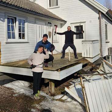 Tres personas derriban alegremente un viejo porche de madera adosado a una casa blanca. La luz del sol ilumina los montones de nieve y las tablas rotas esparcidas por el suelo a su alrededor.