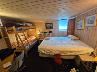 Cette chambre en bois, très chaleureuse, comprend un lit double, un lit superposé, un petit bureau, une chaise, une commode, des tableaux accrochés aux murs, une lampe et une fenêtre laissant entrer la lumière naturelle.
