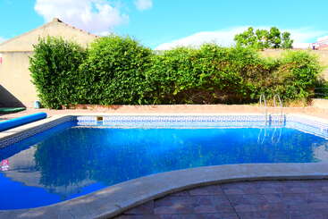 Una piscina azul brillante de agua cristalina se encuentra en un patio trasero embaldosado, rodeada de arbustos verdes y un muro de hormigón bajo un cielo soleado.
