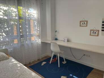 Una habitación luminosa con una gran ventana, cortinas transparentes, una cama, un largo escritorio blanco, una silla blanca, una alfombra azul y dos dibujos enmarcados en la pared.