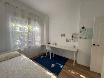 Dormitorio luminoso y ordenado con cortinas transparentes, una cama sencilla, un largo escritorio blanco, una silla blanca, adornos en la pared y una alfombra azul sobre suelo de madera. Estilo minimalista.