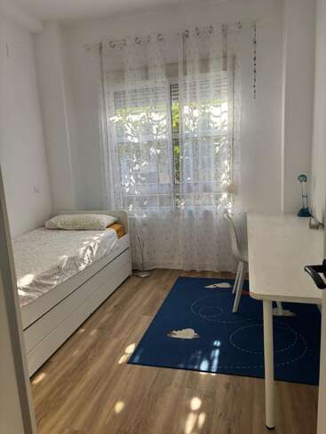 Un dormitorio luminoso y ordenado con una cama individual, muebles blancos, una alfombra azul con nubes, cortinas transparentes, suelo de madera, escritorio, silla y luz solar natural que entra a raudales.