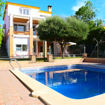 A modern, three-story house with yellow roof tiles, balconies, shaded patios, lush green trees, and a beautiful backyard swimming pool under a clear blue sky.
