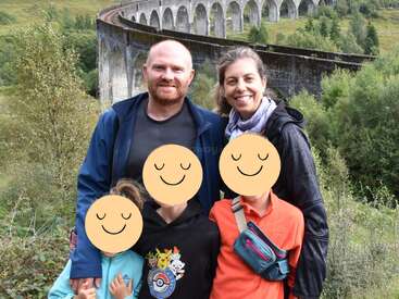 Una familia de cuatro miembros posa al aire libre cerca de un gran viaducto de piedra rodeado de exuberante vegetación. Las caras de los niños están cubiertas con iconos de caras sonrientes.