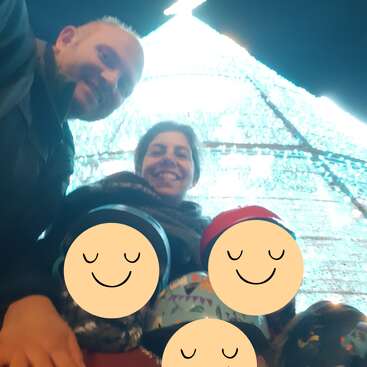 A smiling family poses together in front of a brightly lit, towering Christmas tree. Children's faces are covered with smiley emojis, creating a cheerful, festive atmosphere.