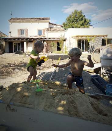 Two young children play joyfully in a large sandbox, using toy trucks and shovels, with a house and trees in the sunny background, enjoying summer fun.