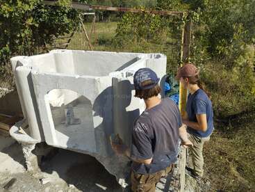 Two people are working outdoors, constructing or plastering a large, white, hexagonal or octagonal cement structure, possibly an oven, amid greenery and sunlight.