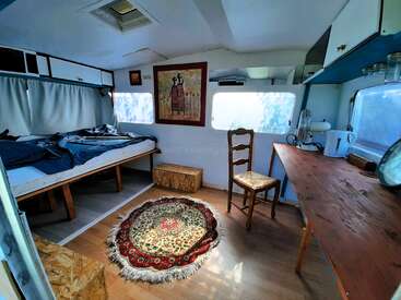 This image shows a cozy, sunlit camper interior with a bed, wooden table, chair, patterned rug, kitchen appliances, artwork on the wall, and storage cabinets.