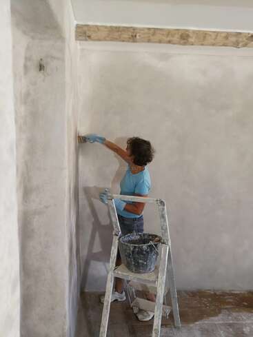 A person wearing blue gloves and a blue shirt is painting or plastering a wall, standing on the floor beside a bucket on a stepladder. Room appears unfinished.