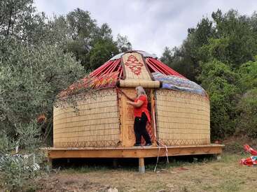 A woman is assembling or decorating a traditional yurt made of wood and woven materials, surrounded by trees and greenery, under a cloudy sky in nature.