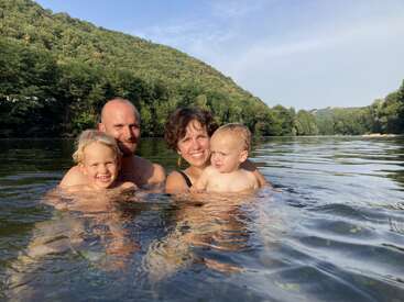 A joyful family of four is swimming together in a scenic river with lush green hills in the background, enjoying a sunny, relaxing day outdoors.