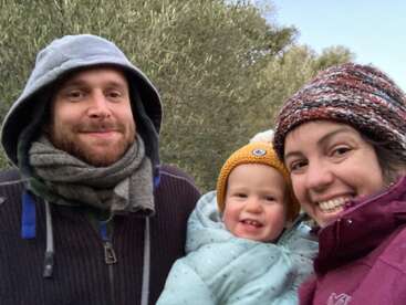 The image shows a family of three, consisting of a man, a woman, and a baby, posing for a selfie in a wooded area on a chilly day.
