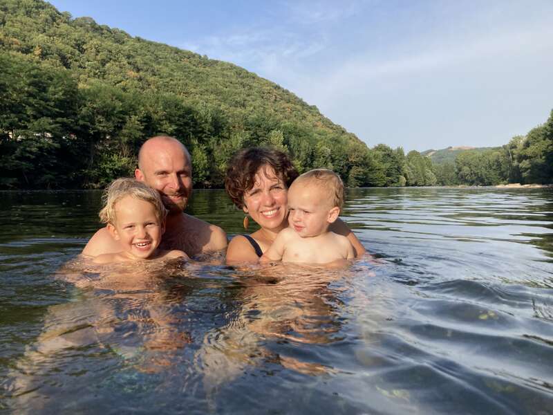A joyful family of four is swimming together in a scenic river with lush green hills in the background, enjoying a sunny, relaxing day outdoors.
