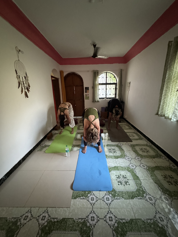 Drei Personen üben drinnen Yoga, indem sie auf Matten in einer Vorwärtsbeuge stehen. Der Raum hat einen gemusterten Fliesenboden, Fenster mit Vorhängen und einen Traumfänger an der Wand.