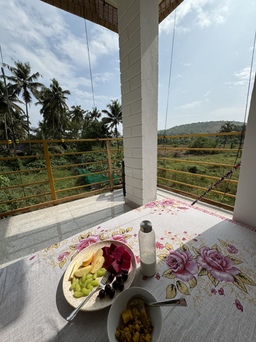 Ein Frühstückstisch mit frischem Obst, einer Schüssel Curry und einer Flasche Milch steht auf einem Blumentuch, mit Blick auf tropisches Grün und ferne Hügel.