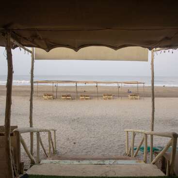 This image shows a view from a shaded beach hut looking out onto a sandy beach with wooden loungers and umbrellas, and the ocean in the distance.