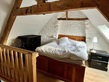 Um quarto aconchegante no sótão com vigas de madeira à vista, paredes de pedra caiadas de branco, uma cama de madeira, cômodas escuras, abajur, claraboia e uma grade de madeira. Um ambiente convidativo.