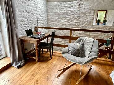 Este aconchegante espaço de trabalho em loft conta com uma mesa rústica de madeira com um laptop, uma cadeira preta, uma cadeira de balanço almofadada e paredes brancas texturizadas, transmitindo conforto e aconchego.
