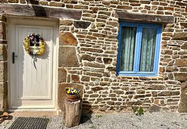 Uma casinha rústica de pedra tem uma porta branca com uma guirlanda de flores, uma janela com moldura azul e cortinas de renda, e um vaso de planta apoiado em um toco de árvore.