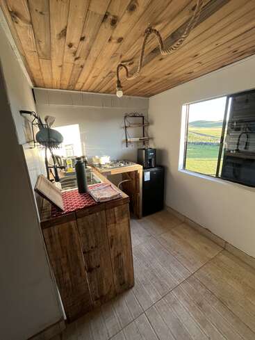 Cette cuisine confortable est équipée d'armoires rustiques en bois, d'un plafond en bois, d'un petit réfrigérateur, d'étagères ouvertes, d'un éclairage simple et d'une fenêtre offrant une vue panoramique sur la verdure.