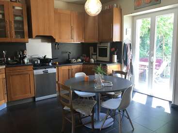 A cozy kitchen with wooden cabinets, round dining table, assorted chairs, appliances, and utensils. Sunlight streams through glass doors leading to a green outdoor area.