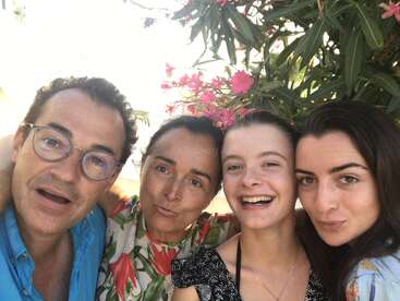 A happy family of four poses for a selfie outdoors, with bright sunlight, smiles, and pink flowers from a bush providing a cheerful, vibrant background.