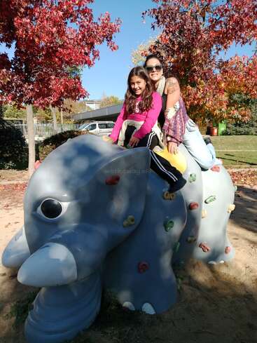 Una mujer y una niña se sientan felices en una gran escultura gris con forma de elefante en un parque infantil. Las hojas otoñales, el sol brillante y las sonrisas crean una alegre escena al aire libre en el parque.