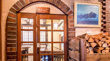 La imagen muestra un interior rústico con puertas de madera bajo un arco de ladrillo, leña apilada, un cuadro de montaña y letreros japoneses, que crean un ambiente acogedor y cálido.