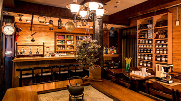 Esta acogedora cafetería cuenta con paredes de madera, farolillos vintage y estanterías repletas de jarras y té, un hogar central con tetera, una cálida iluminación y acogedores asientos en el mostrador.