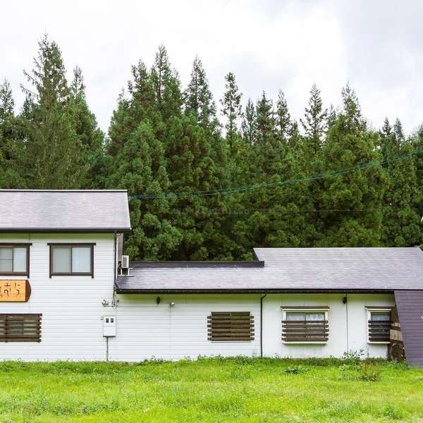Una casa blanca de dos plantas con adornos oscuros se asienta en un patio verde cubierto de hierba, rodeada de densos árboles y bosque. Sobre la puerta cuelga un cartel de madera.