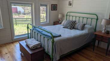 A cozy bedroom features a green metal bed frame, two windows, wooden floors, nightstands with lamps, a trunk, simple decor, and views of a peaceful farm.