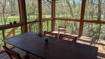 A cozy screened porch with a wooden table, four chairs, and a vase. Sunlight streams in, overlooking a green yard surrounded by trees and nature.