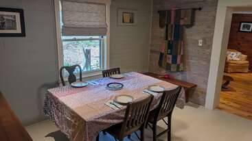 A cozy dining area with a square table set for four, featuring a patterned tablecloth, wooden chairs, wall art, quilt decor, and a window view.