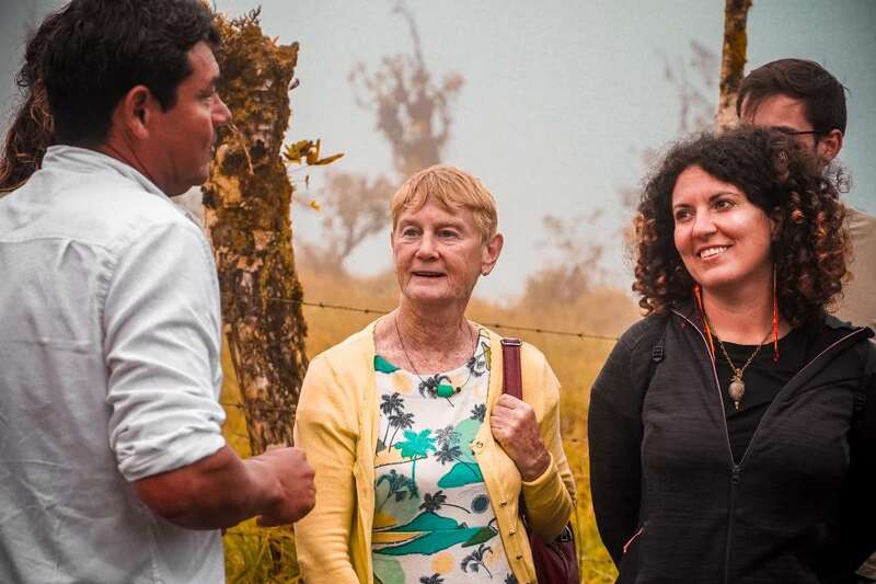 Four people stand outdoors, engaged in conversation. The background is misty, with trees and a fence. Everyone appears interested, with two women smiling warmly and listening attentively.