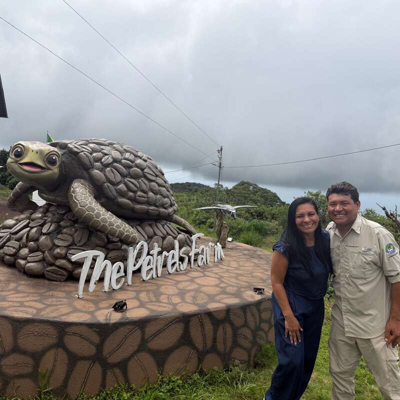 Two people stand closely together outdoors, smiling happily. The background includes lush greenery, cloudy skies, and a rustic setting, suggesting they are enjoying a nature outing.