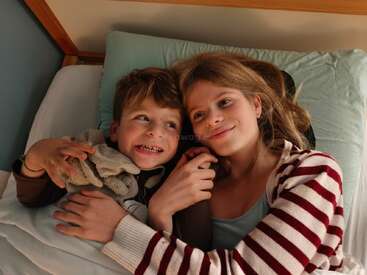 Un petit garçon et une petite fille sont allongés dans leur lit, blottis sous une couverture. Ils s'échangent des sourires chaleureux, rayonnant d'amour, de réconfort et d'affection fraternelle. Une atmosphère chaleureuse.