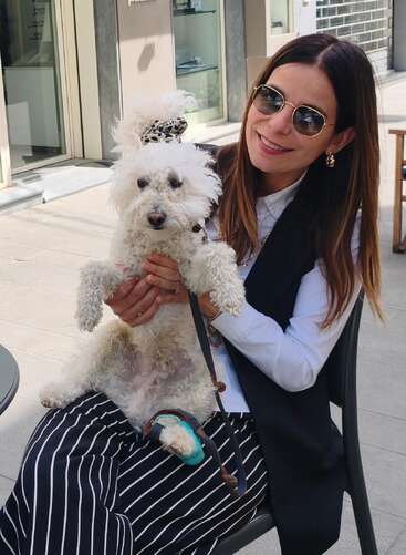 Une femme souriante, portant des lunettes de soleil, tient dans ses bras un chien blanc tout doux qui porte un élastique à cheveux à imprimé léopard. Ils sont assis à la terrasse d'un café par une journée ensoleillée.