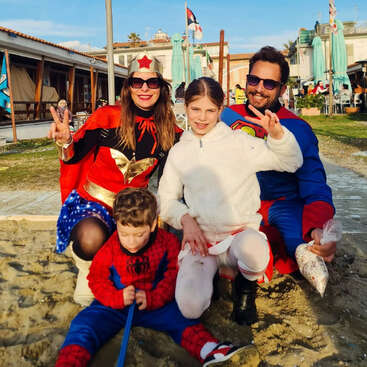 Une famille déguisée en super-héros — Wonder Woman, Superman et Spider-Man — pose joyeusement sur une plage de sable, en compagnie de deux enfants, profitant ensemble d'une journée ensoleillée.