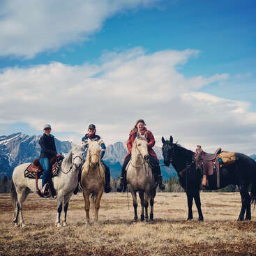 The image depicts three individuals riding horses in a field with mountains in the background, all wearing warm clothing and smiling at the camera.