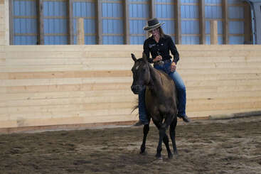 A woman in a cowboy hat rides a brown horse inside a wooden indoor arena, guiding it skillfully along the sandy ground, demonstrating horsemanship.