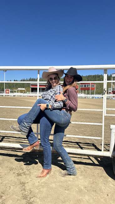 Two women dressed in western attire and cowboy hats pose playfully in a rodeo arena, one lifting the other. Bright sunny day, blue sky, joyful atmosphere.