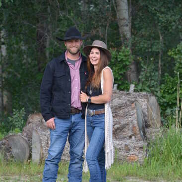 A smiling couple stands outdoors, both wearing hats, jeans, and casual Western attire. They're surrounded by trees and logs, radiating happiness and warmth.