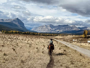 A person rides a horse along a dirt path in an expansive, dry field, surrounded by mountains, cloudy skies, and scattered trees with autumn foliage.