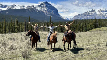Three people on horseback raise their arms joyfully in a scenic mountain landscape, surrounded by green pine trees, grassy field, blue sky, and snow-capped peaks.