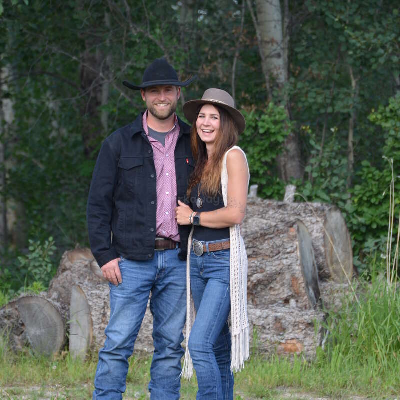 A smiling couple stands outdoors, both wearing hats, jeans, and casual Western attire. They're surrounded by trees and logs, radiating happiness and warmth.