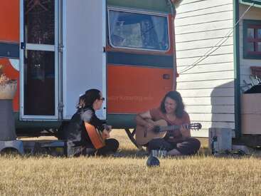 Duas mulheres sentam de pernas cruzadas na grama seca, tocando violões juntas do lado de fora de uma caravana e uma casinha, curtindo a música e o clima ensolarado e relaxante ao ar livre.