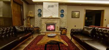 This cozy living room features two dark leather sofas, a crackling fireplace, a unique wooden coffee table, decorative plates, warm lighting, and a vibrant red rug.