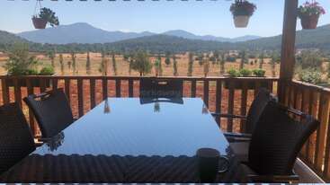 This image shows a patio table with four chairs and a coffee mug, overlooking a scenic countryside with mountains, trees, fields, and hanging flower pots.
